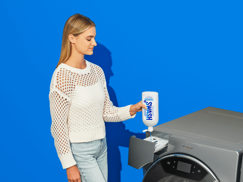 A woman dispensing Swash laundry detergent directly into a top loading washing machine using the Precision Pour Cap