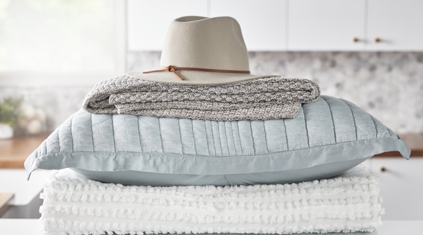 A stack of blankets, pillows and a hat sit on a laundry room counter.