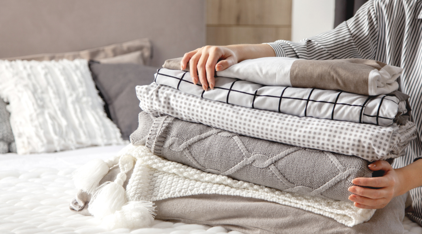 A person holds a stack of fitted sheets and blankets on top of a bed.